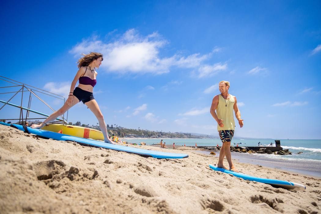 A student stepping onto a surfboard on the sand while Troy MacDonald watches